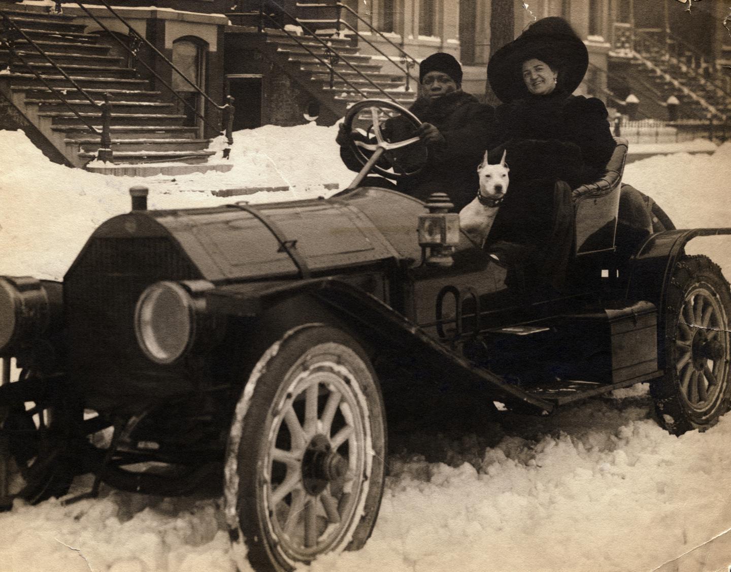 Jack Johnson sits next to his wife, who wears a large hat, in a car that is parked in the snow. They are accompanied by a dog.