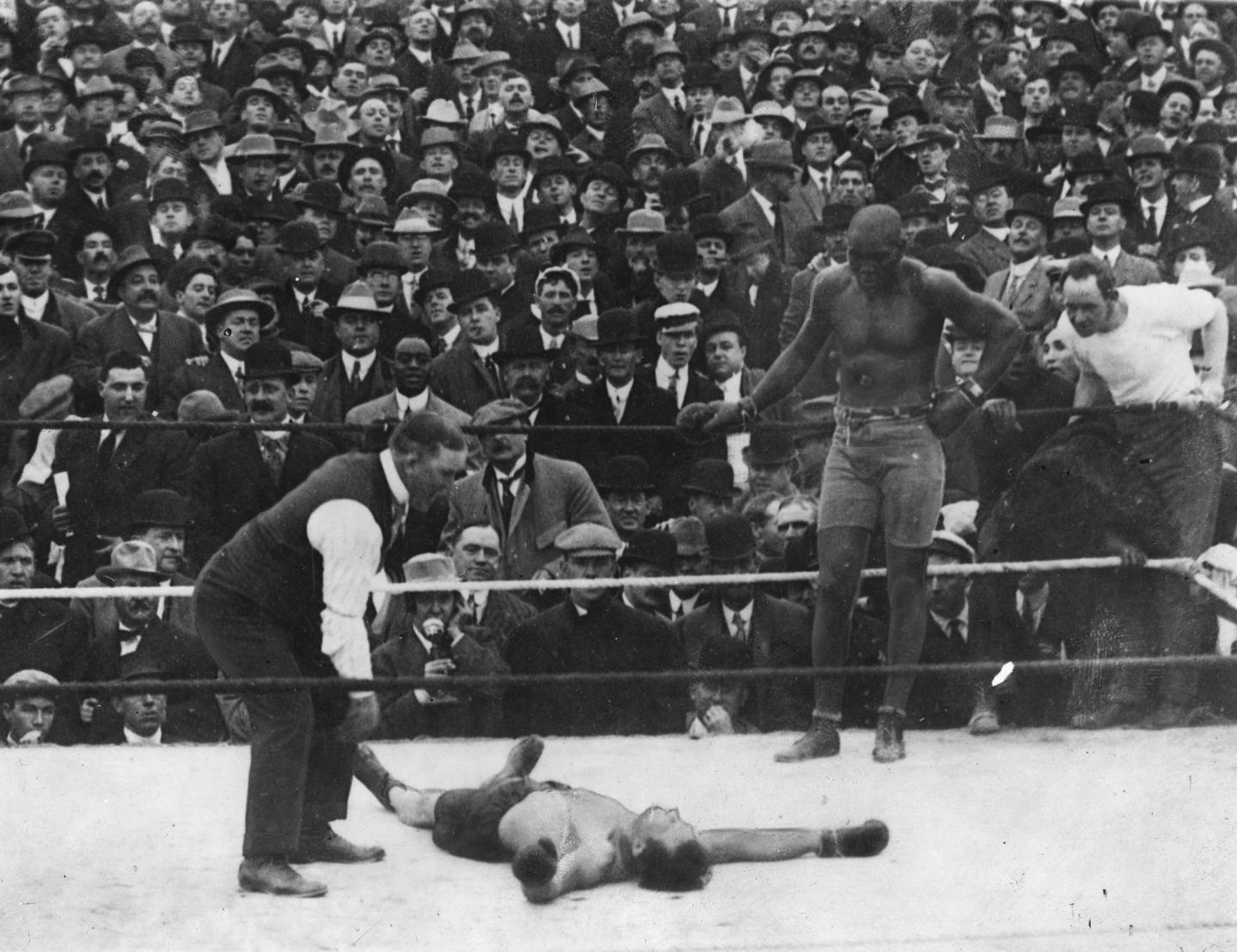 Jack Johnson leans back on the ropes while his opponent, Stan Ketchel, lays unconscious in the ring and the referee bends over him to count him out.