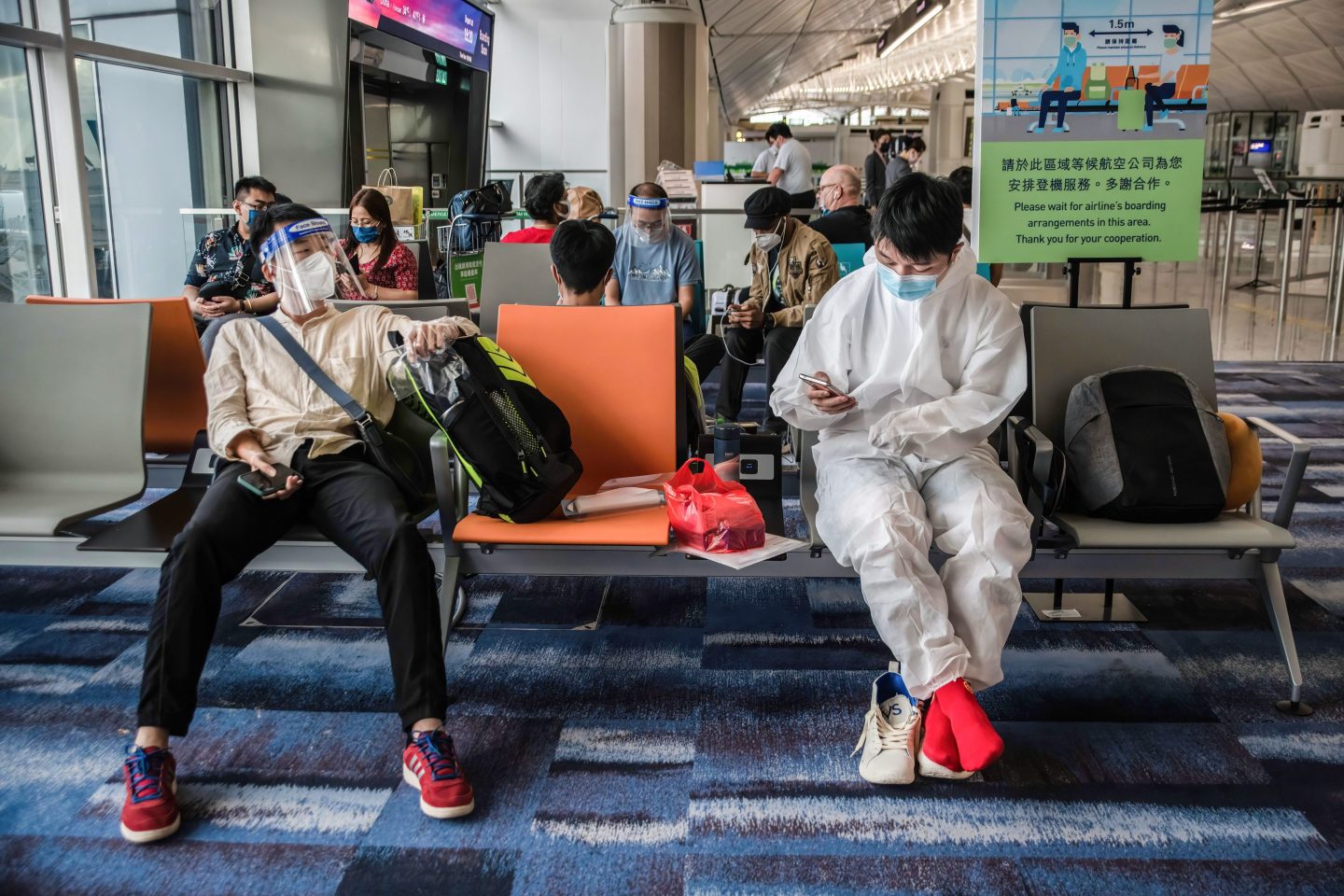 Two passengers wait at Hong Kong airport, with one wearing personal protective equipment