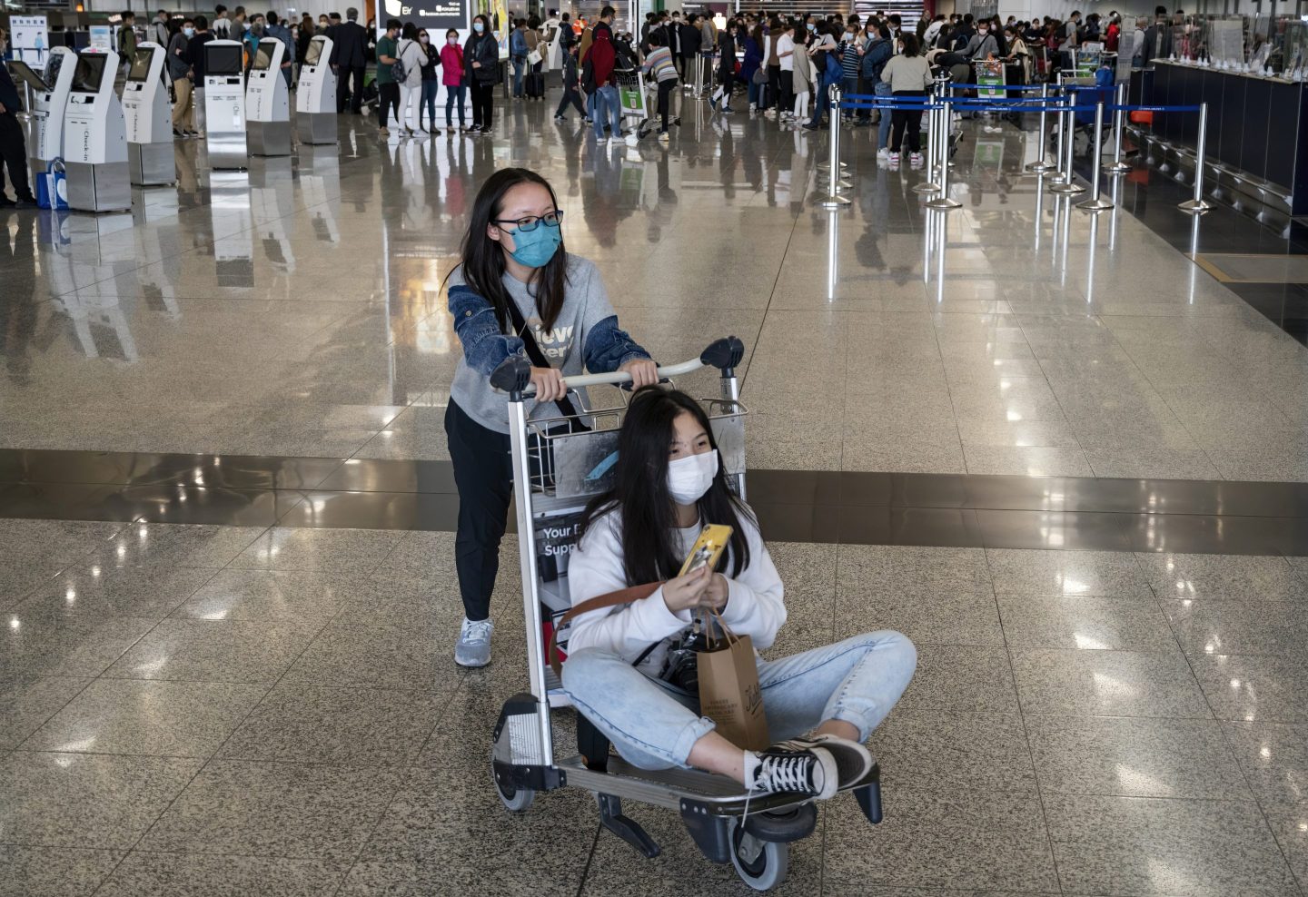 Woman pushes friend in luggage cart at Hong Kong airport.
