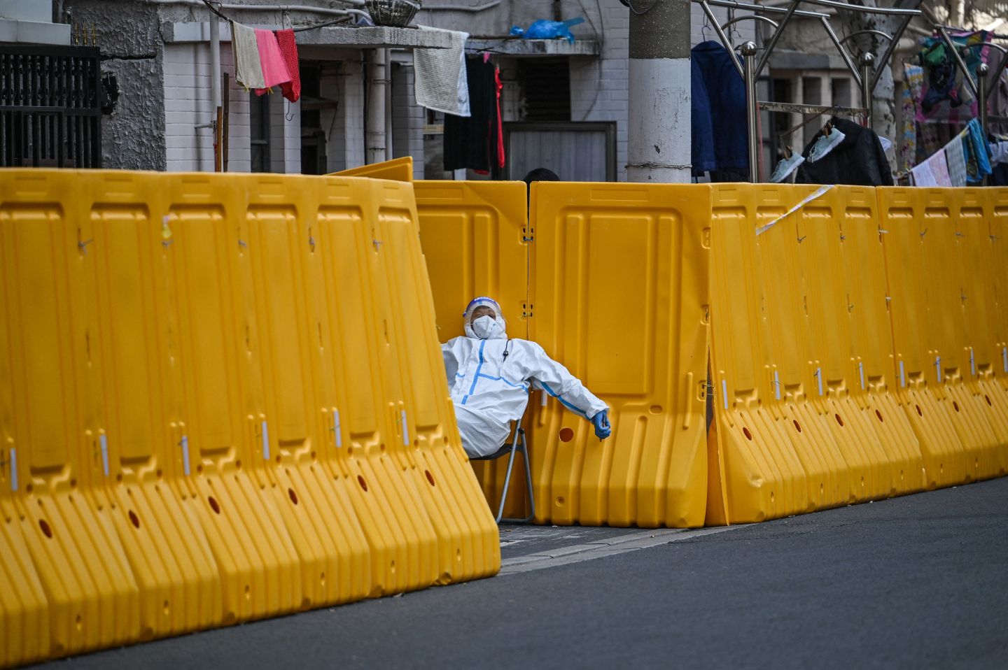 A worker in a hazmat suit naps while guarding the entrance to a building blocked by yellow barricades