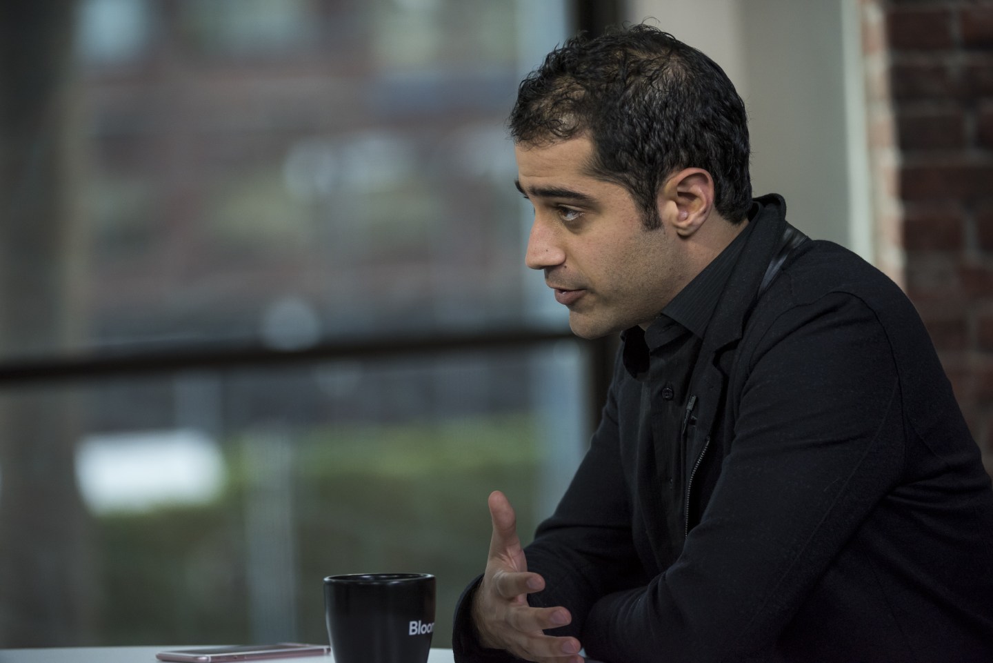 Kayvon Beykpour is seen speaking to a reporter (who is off-screen) during an interview with Bloomberg. Beykpour's phone and a cup of coffee lay on the table in front of him.