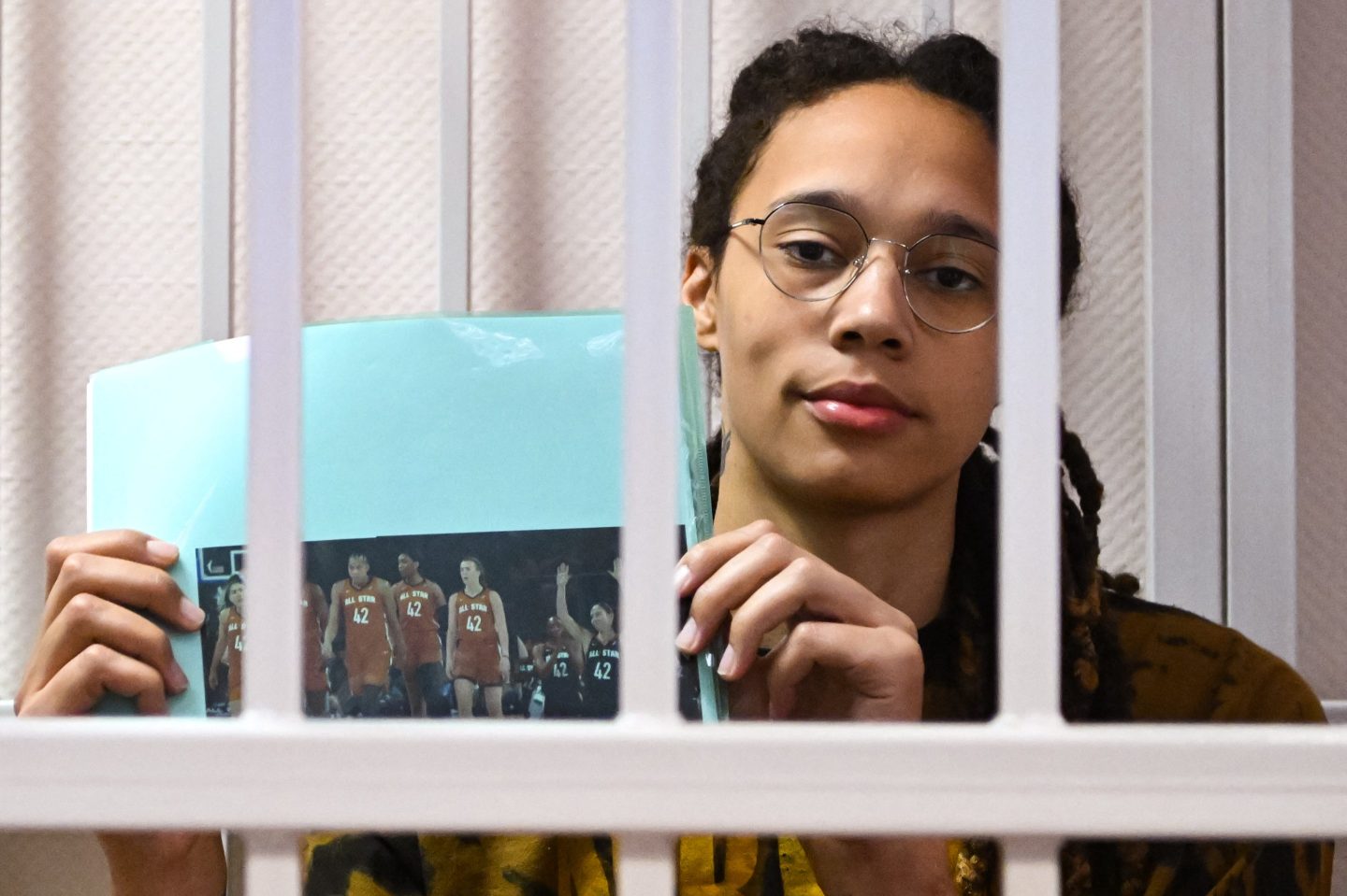 Brittney Griner holds a picture of her WNBA colleagues playing on the court wearing her jersey while she sits inside a cell.