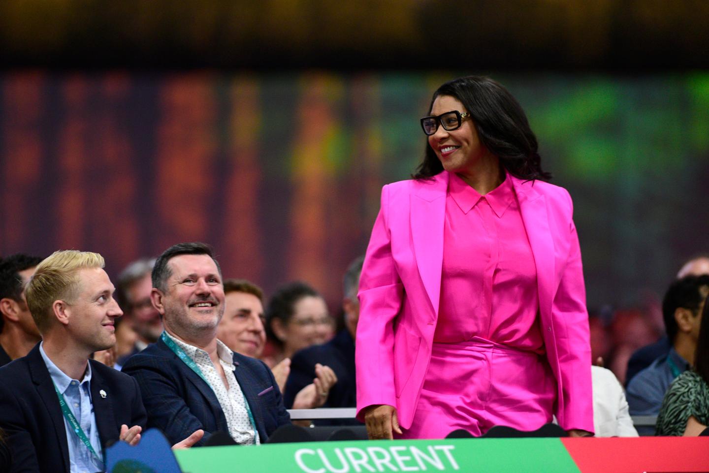 London Breed, mayor of San Francisco, during the 2022 Dreamforce conference in San Francisco, wearing a fully bright pink suit, thick framed black glasses, smiling at the crowd as she stands up.