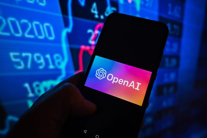 A mobile phone screen with the OpenAI logo in front of a large monitor displaying financial data