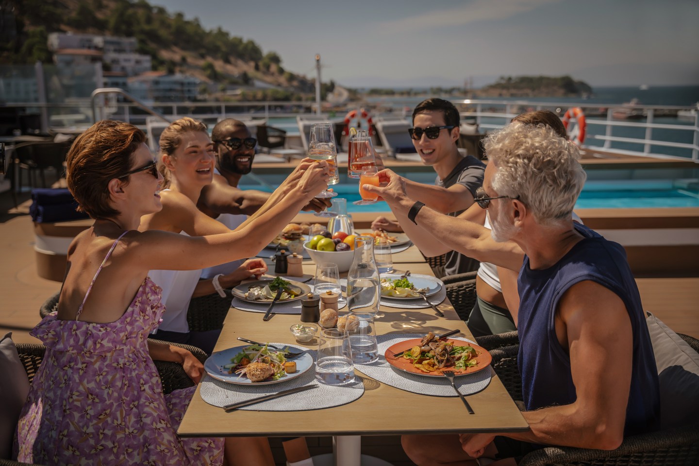 A group of adults holding drinks to cheers while eating a meal on a boat at sea.