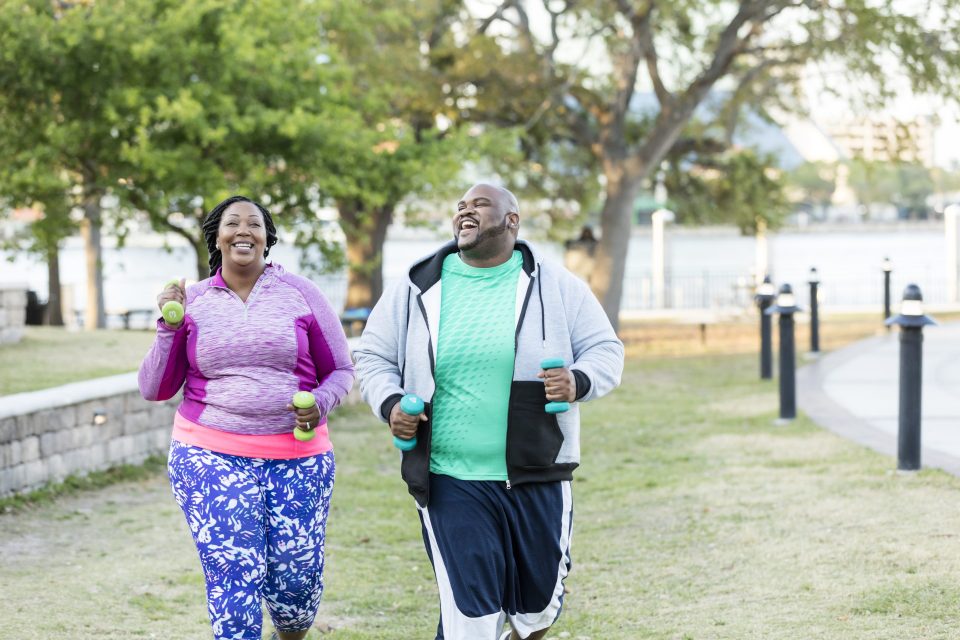 An African-American couple in the park, exercising together in a city park, conversing as they power walk with hand weights. They are both plus size models with large builds.